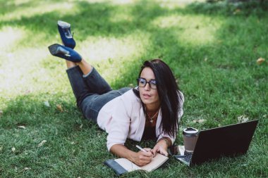 İş kadını, yaz çimenleri parkında dizüstü bilgisayar kullanarak yatıyor. Karantina sırasında uzaktan kumanda. Kahveli kadın. Açık havada. Yumuşak seçici odak.