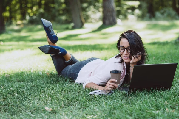 女商人躺在一个夏季草园里 手里拿着笔记本电脑 手里拿着一杯咖啡 隔离期间的远程工作 喝咖啡的女人户外 软性选择性重点 — 图库照片