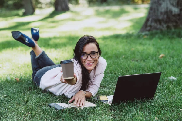 女商人躺在一个夏日草园里 用笔记本电脑微笑着 端着一杯咖啡 隔离期间的远程工作 喝咖啡的女人户外 软性选择性重点 — 图库照片