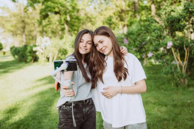 Gülümseyen genç kadın etkili blog yazarları dışarıda güneşli yeşil bir parkta akıllı fonlarıyla bir dengeleyiciyle video kaydediyorlar. Blog konsepti. Yumuşak seçici odak.