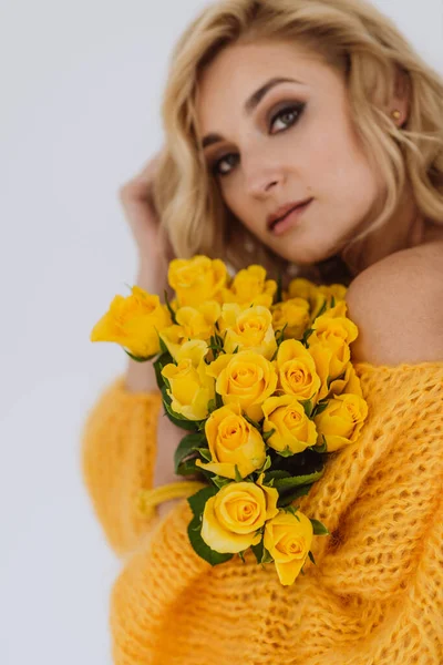 スタジオの白い背景にボリュームのあるセーターに黄色のバラの花束を持つ美しい若いブロンドの女性の肖像画 柔らかい選択的フォーカス — ストック写真