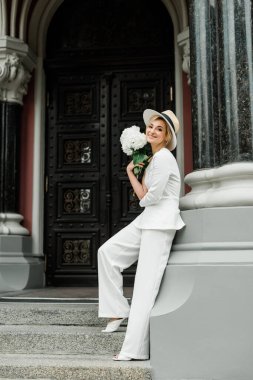Beyaz pantolonlu şık bir bayan ve şehrin sokaklarında bir buket ortanca çiçekli zarif bir saç stili olan bir şapka. Gelin olan kadın. Yumuşak seçici odak
