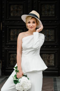 Beyaz pantolonlu şık bir bayan ve şehrin sokaklarında bir buket ortanca çiçekli zarif bir saç stili olan bir şapka. Gelin olan kadındır..
