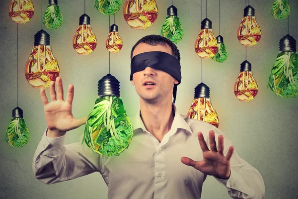 Blindfolded man walking through light bulbs shaped as junk food green vegetables — Stock Photo, Image