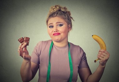 Confused looking woman with chocolate and banana trying to make healthy choice  clipart