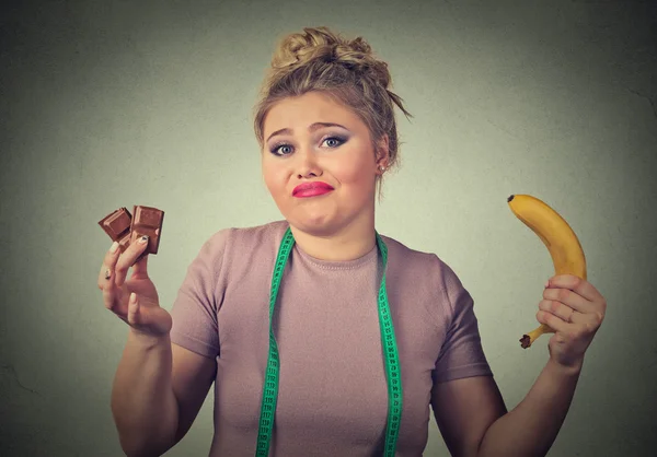 Mulher de aparência confusa com chocolate e banana tentando fazer uma escolha saudável — Fotografia de Stock