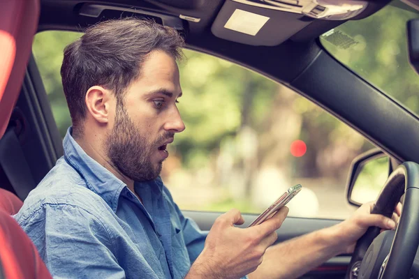 Hombre sentado en el coche con el teléfono móvil en los mensajes de texto de la mano mientras conduce — Foto de Stock