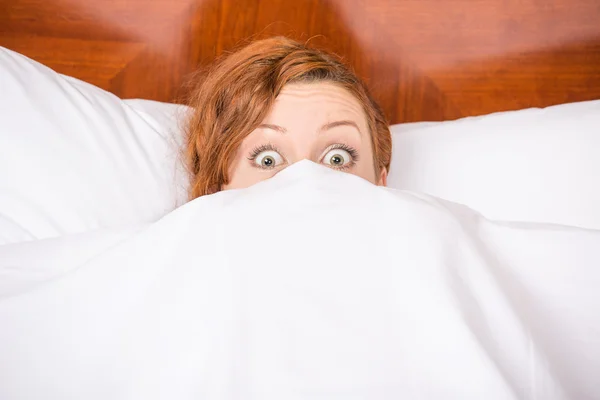 Funny young brunette woman in bed looking and peeking over sheets surprised — Stock Photo, Image