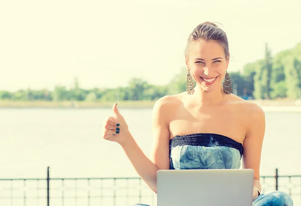 Lachende vrouw aan zee zittend op gras met haar computer gebalanceerd op knieën — Stockfoto