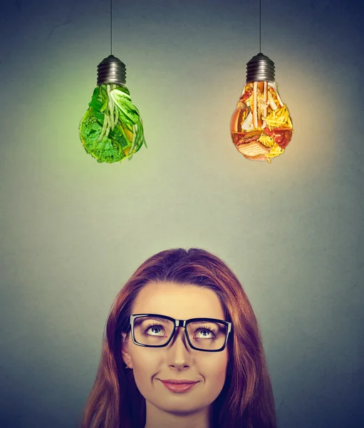 Женщина думает, глядя вверх на нездоровую пищу и зеленые овощи лампочки — стоковое фото