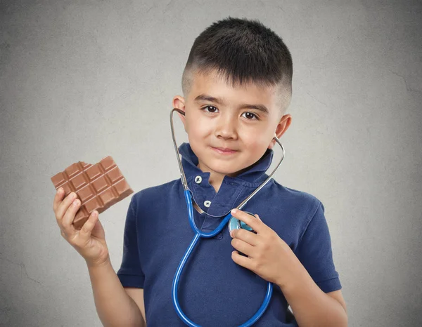 Menino comendo chocolate ouvindo seu coração com estetoscópio — Fotografia de Stock