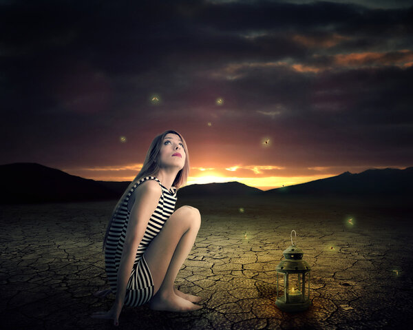 Lonely woman with antique light lamp sitting in a middle of a desert looking up on night skyline falling stars with mountains background. Dreamland imagination screen saver. Human expression emotions