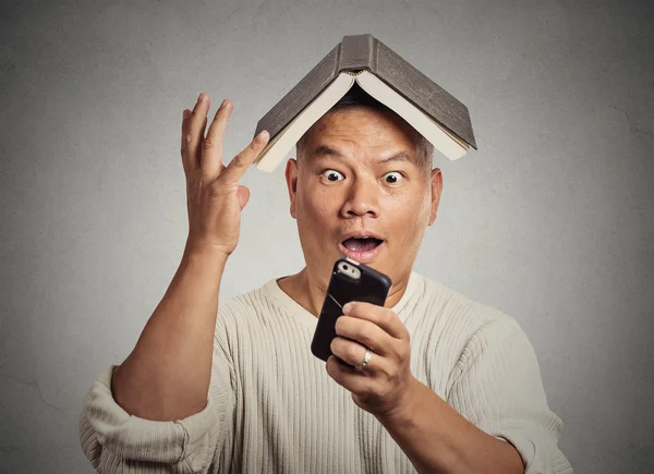 Surprised funny looking man with book on head reading news on smartphone — Stock Photo, Image