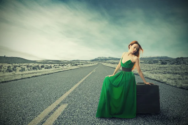 Женщина, сидящая на чемодане на сельской дороге в ожидании поездки — стоковое фото