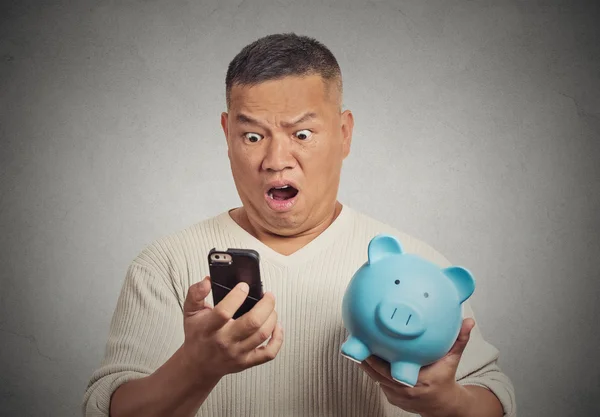 Homem chocado olhando para seu telefone inteligente segurando porquinho banco — Fotografia de Stock