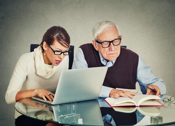 Жінка працює на ноутбуці і старший дідусь чоловік читає з книги — стокове фото