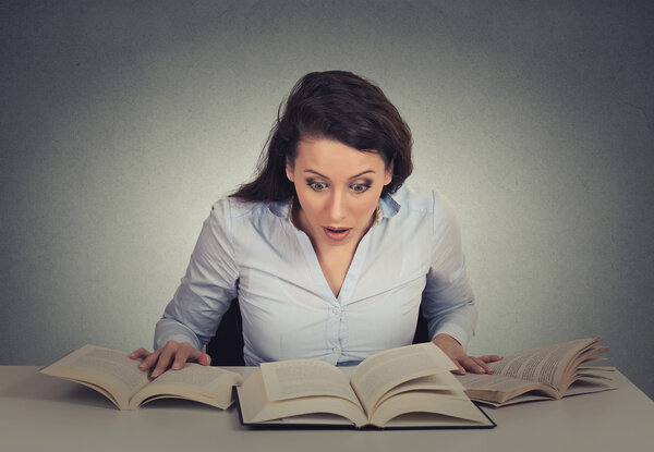 shocked woman sitting at desk with many opened books reading 