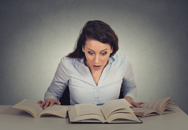 shocked woman sitting at desk with many opened books reading 