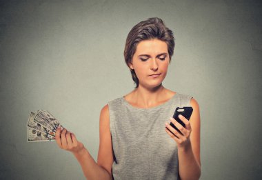 Unhappy young woman looking at smart phone throwing away cash dollar bills