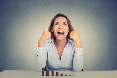 happy woman sitting at table with growing stack of coins showing thumbs up