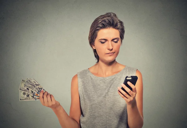 Unhappy young woman looking at smart phone throwing away cash dollar bills — Zdjęcie stockowe