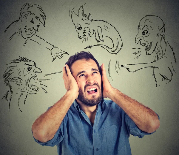 Stressed young man covers his ears with his hands evil guys pointing fingers at him — Stockfoto