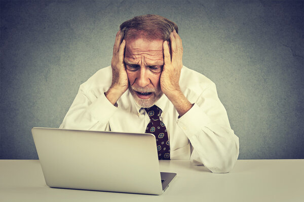 senior stressed man working on laptop sitting at table isolated on gray wall background 