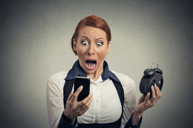 surprised business woman with alarm clock looking at smart phone 