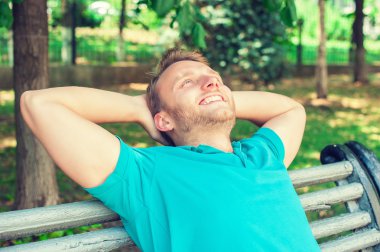 happy handsome young man in shirt looking upwards in thought, relaxing on a bench