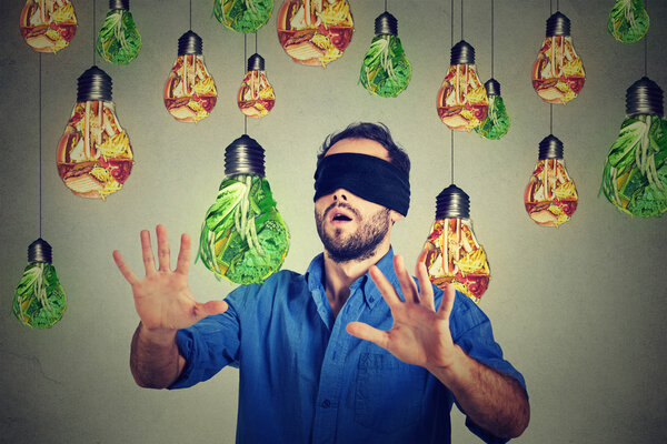 Blindfolded man walking through light bulbs shaped as junk food and green vegetables
