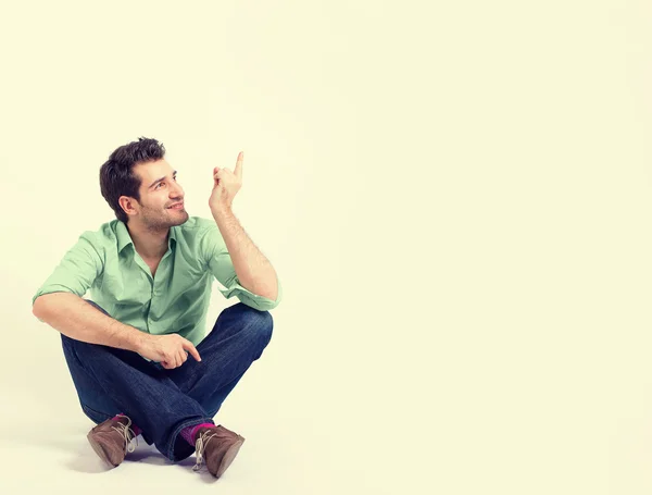 Happy man in green shirt and blue jeans sitting on the floor pointing at blank copy space — Stockfoto