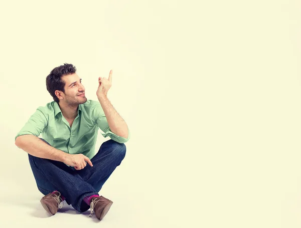 Hombre feliz con camisa verde y vaqueros azules sentado en el suelo apuntando al espacio de copia en blanco — Foto de Stock