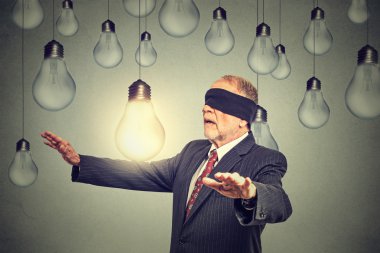 Blindfolded senior man walking through light bulbs searching for bright idea 