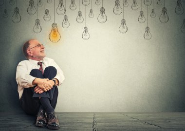 Thinking elderly man in glasses sitting on floor looking up with light idea bulb above head 