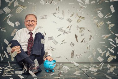 Excited happy senior man sitting on a floor with piggy bank under a money rain 