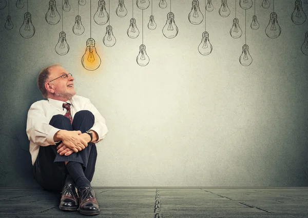 Thinking elderly man in glasses sitting on floor looking up with light idea bulb above head — Stock Photo, Image