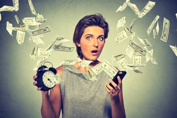 Surprised woman with alarm clock smart phone and dollar banknotes flying away. Online income business concept — Stockfoto