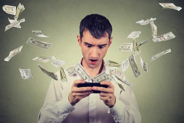 Shocked young man using smartphone with dollar bills banknotes flying away — Stock Photo, Image