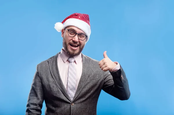 Gri takım elbiseli, beyaz sakallı, pembe tişörtlü, kravat çizgili Noel Baba gülüşlü genç adam açık mavi arka planda mutlu görünüyor. Noel tatili tatili konsepti. Boş metin kopyalama alanı