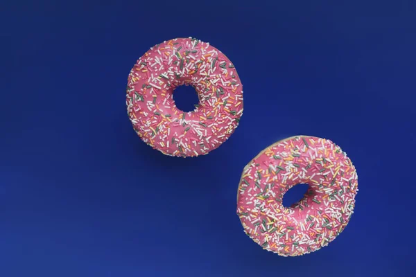 Two pink glazed donuts with colourful sprinlkes falling floating or flying isolated in dark blue background. Levitating food bakery sweet tasty dessert side view