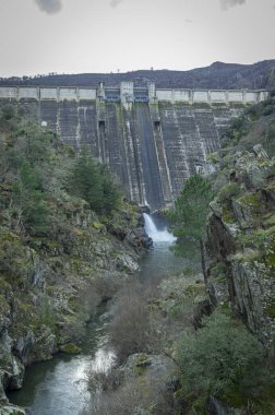 Yıpranmış bir hidroelektrik barajındaki su boşaltma borusunun yakın plan detayları. Beton duvar, koyu su lekeleri, yosun büyümesi ve aşınma ile birlikte ciddi yaşlanma belirtileri gösteriyor. Altyapı bakım sorunlarını resmediyor