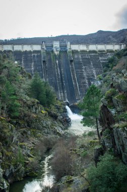 Büyük beton hidroelektrik baraj kayalık bir nehir yatağına su akıntısı salıyor. Altyapı, çam ağaçlarıyla çevrili vahşi bir dağ manzarasıyla çevrilidir. Yenilenebilir enerji kavramı, su kaynağı yönetimi ve inşaat mühendisliği