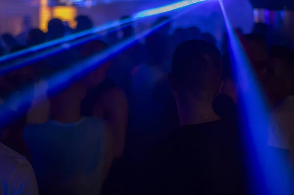 Kalabalık bir dans pistinin atmosferik çekimi bir gece kulübünde. Canlı mavi lazer ışınları ve neon ışıkları altında eğlenen genç insanların siluetleri. Gece hayatı, eğlence ve gençlik kültürü için mükemmel.