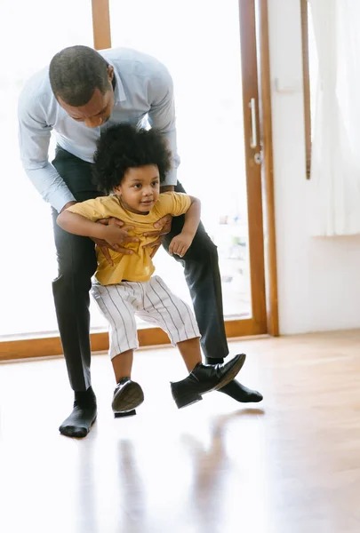 幸せなアフリカ系アメリカ人の父親は 子供が仕事の後に父親の靴で遊んでいる間に家に小さな息子を運びます お帰りなさいパパや家族の再会のコンセプト — ストック写真