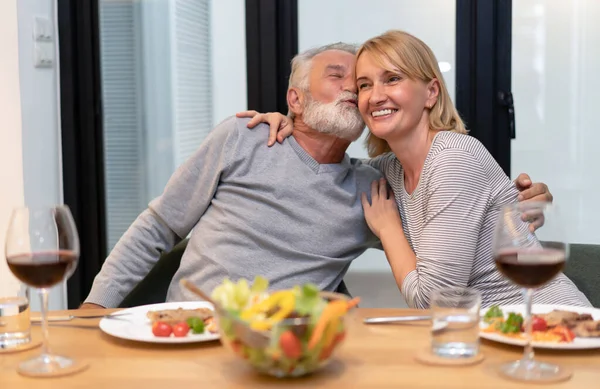 Cheerful senior couple embracing in love hug and kiss eating salad and drinking red wine together with healthy food on the kitchen at home. Concept of healthy nutrition in older age and Happy retirement.