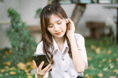 Üniversite kampüsünün yanındaki parkta oturmuş kulaklıkla favori şarkıları dinleyen üniformalı genç Asyalı öğrenci portresi. Teknoloji ve İletişim Konsepti.