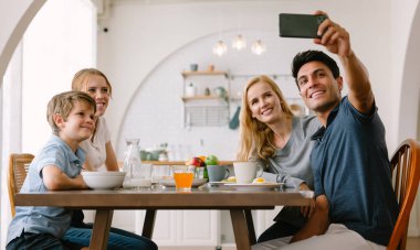 Mutlu aile portresi, evde masada kahvaltı ederken akıllı telefondan selfie için poz veriyor. Aile, anne, baba, kız ve oğuldur. Afiyet olsun. Aile tatili ve birliktelik.