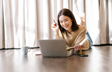 Genç Asyalı güzel bir kadının portresi evde rahat bir oturma odasında modern dizüstü bilgisayar kullanarak çalışarak ve bir deftere fikirler yazarak çalışıyor. Serbest bir iş. Akıllı çalışma.