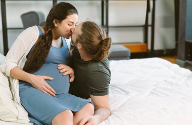 Mutlu hamile çift yatakta dinleniyor ve yatak odasında öpüşüyor. Sevgili çift, Duygu, hamilelik ve annelik.