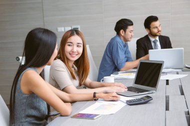 İki Asyalı meslektaşı sırıtırken bir dizüstü bilgisayarda çalışmayı tartışıyor, tablolar ve belgeler masada. Arka planda, iki takım arkadaşı görevlerine ve verimli ofis ortamlarına odaklanıyor.